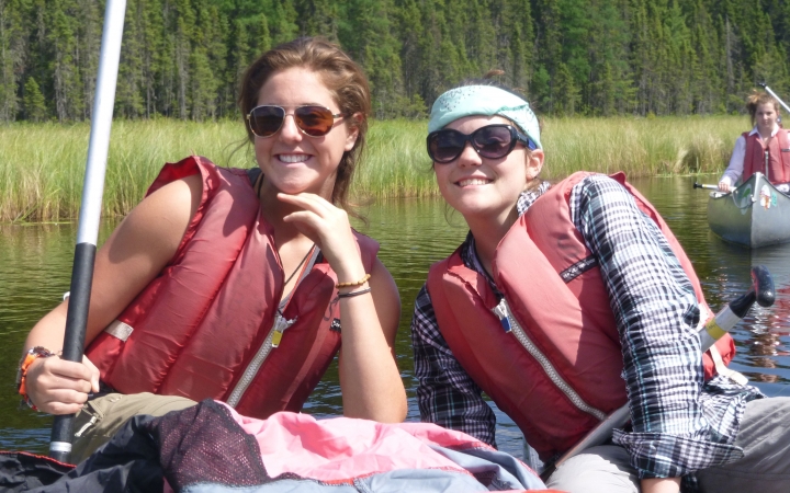 Two students sit in a watercraft and smile at the camera. Behind them is another canoe and the green shore.